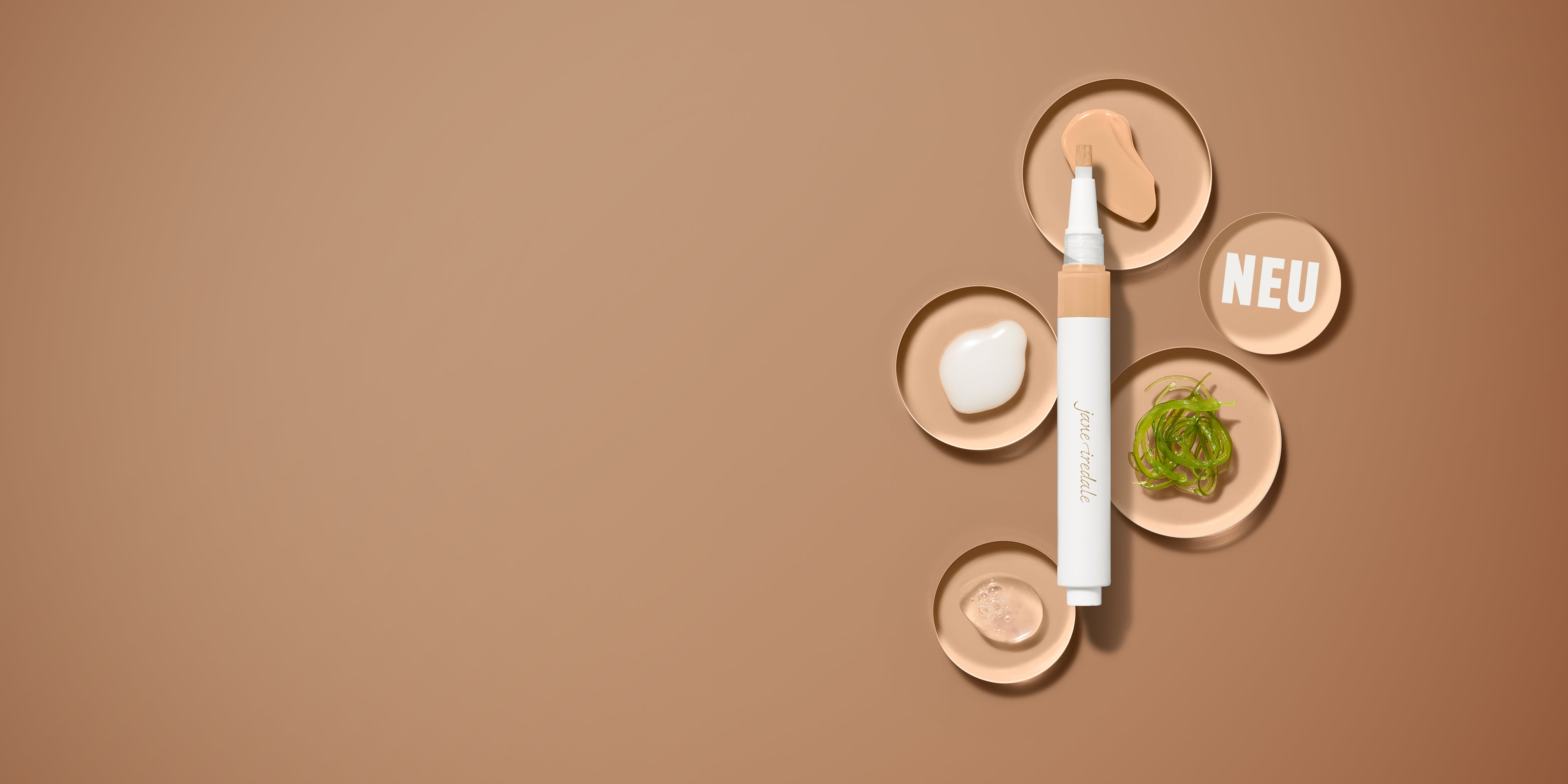 Concealer-Stift der Marke Jane Iredale auf hellbraunem Hintergrund, umgeben von fünf kleinen runden Schälchen. Die Schälchen zeigen verschiedene Texturen und Inhaltsstoffe: weiße Creme, durchsichtiges Gel, grüne Algen und beigefarbene Kosmetikpaste. Ein Schälchen enthält den weißen Schriftzug 'NEU'.